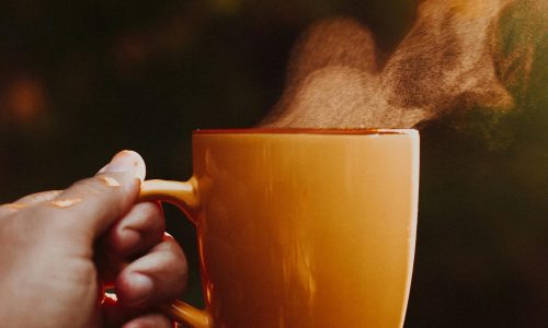 Hand holding a steaming orange mug outdoors in warm morning sunlight, creating a cozy atmosphere.