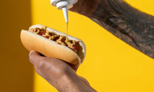 Person applying mustard to a hot dog with fries in the background, on a bright yellow backdrop.